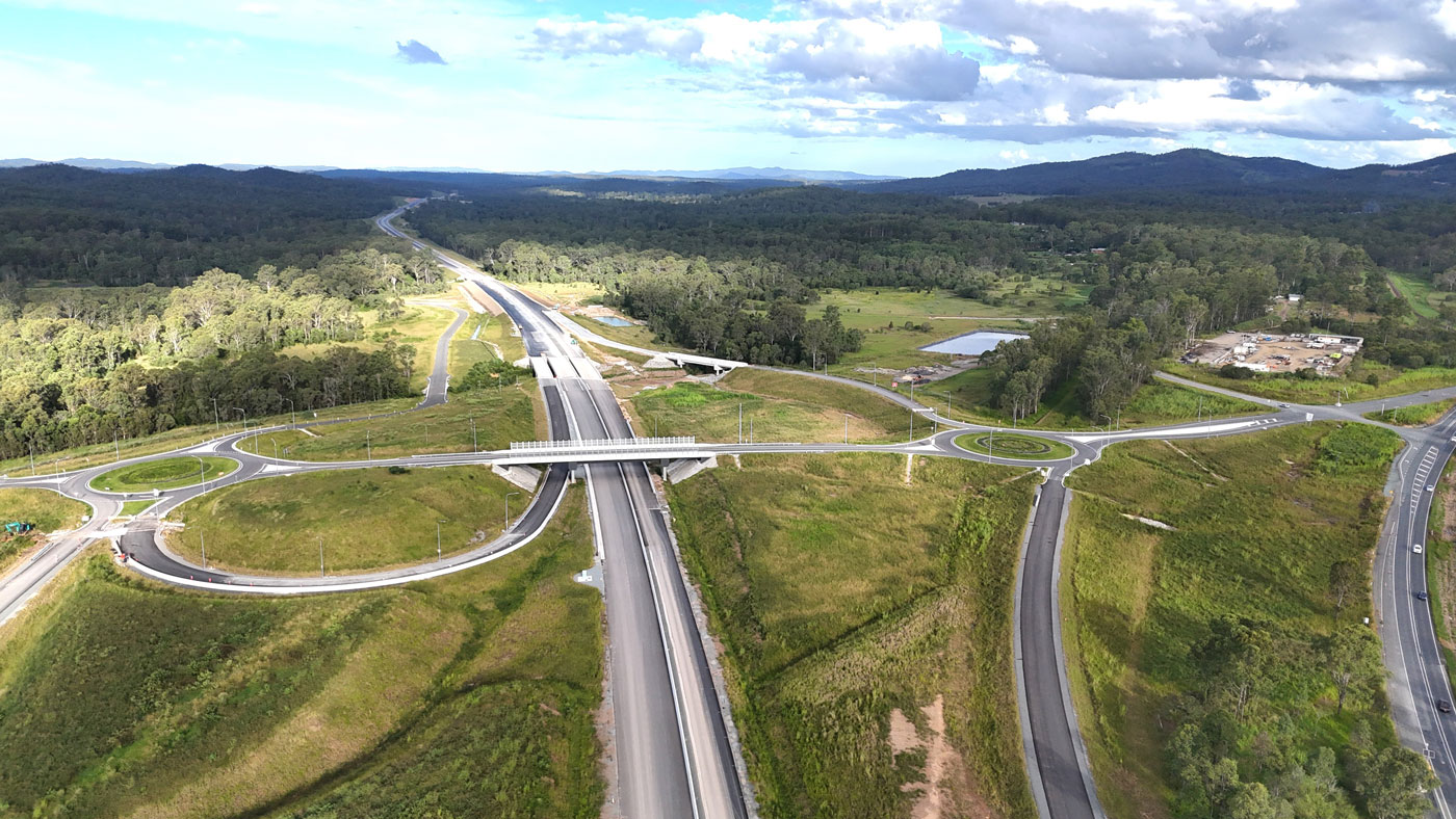 Looking south onto a large interchange with two roundabouts on either side of the new highway running up the centre of image towards small bridges carrying the highway over a small creek. The existing Bruce Highway has traffic travelling on it to the right.
