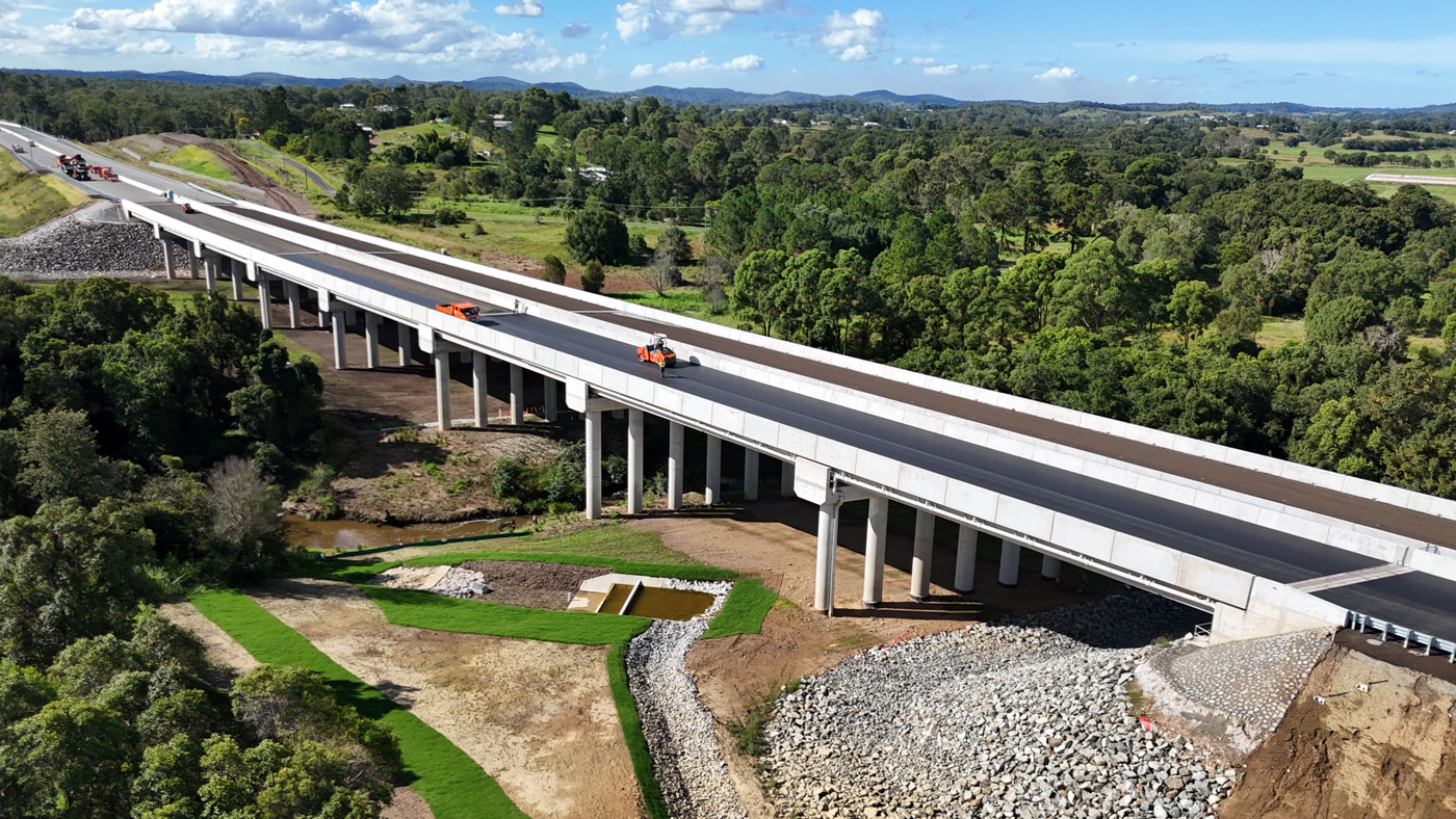 Looking north towards an 8 span, 260 metre long concrete bridge structure over Deep Creek with construction crews on the bridge completing the final layer of pavement in preparation for linemarking. There is a bio-retention basin (like a sediment pond) in the foreground.