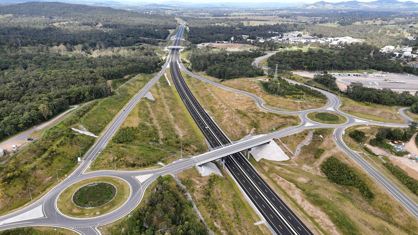 Looking south from two large roundabouts and on/off ramps on either side the new Flood Road interchange onto the new four lane highway with new dark pavement and completed linemarking. Noosa Road bridge traverses over the new highway in the distance.