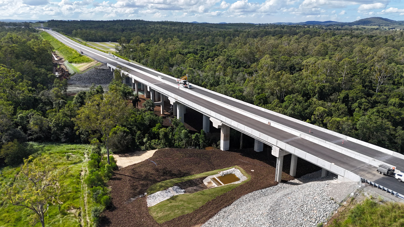 Looking north towards an 8 span, 250 metre long concrete bridge structure over Six Mile Creek with construction crews on the bridge completing pavement and finishing work in preparation for linemarking. There is a bio-retention basin (like a sediment pond) in the foreground.