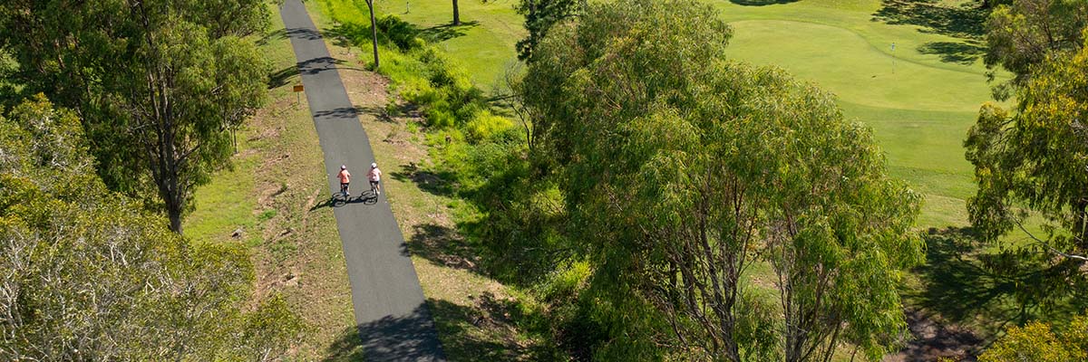 Aerial view of 2 bike riders riding side by side on a path through trees and grassland.