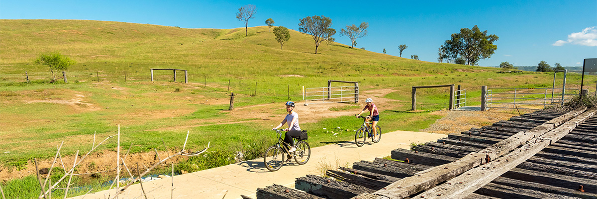 2 bike riders riding on a path between some hilly paddocks and a long row of old wooden slats.