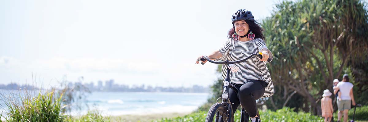 A person riding a bike beside a beach. In the background another person walks with 2 children in the other direction. High rise buildings are visible at the other side of the water. 