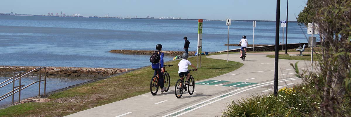 A concrete paths runs alongside a large body of water. 2 bike riders ride side by side and another rides alone further up the path.