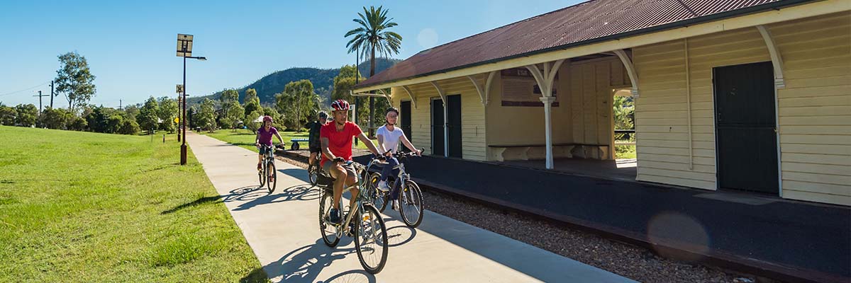 A group of 4 bike riders riding on a concrete path next to an old-style out-of-use train station, with mountains and tropical trees in the background.
