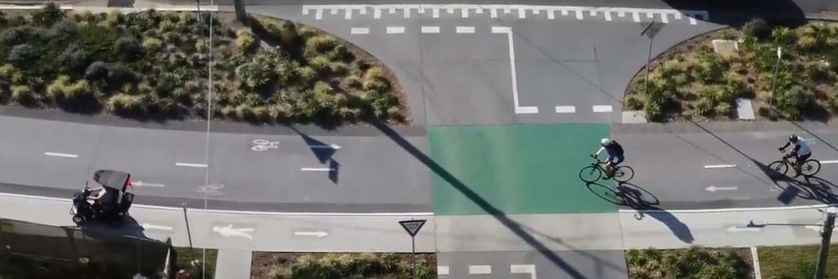 Computer-generated aerial view of parallel bike and pedestrian paths, intersecting with a road. 2 bike riders ride on the bike path and there is a mobility scooter on the pedestrian path. The road has "give way" signs on either side where it intersects with the paths.
