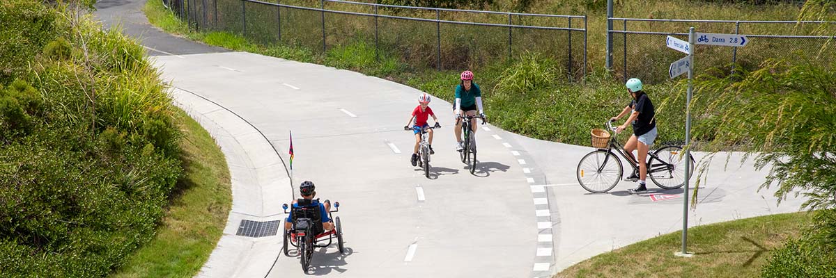 T-intersection between 2 bike paths. An adult and child ride bikes in one direction past the intersection and another person rides a recumbent trike in the other direction. Another person waits on the other path at the intersection.